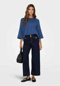 Femme portant un pull bleu ample, des jeans larges taille haute de couleur sombre avec des boutons dorés, des ballerines noires, et tenant un sac à main noir.