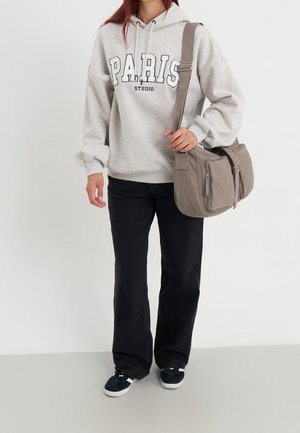 Person trägt einen hellgrauen "PARIS STUDIO" Hoodie, schwarze Hose, schwarz-weiße Sneakers und trägt eine taupefarbene Umhängetasche vor weißem Hintergrund.