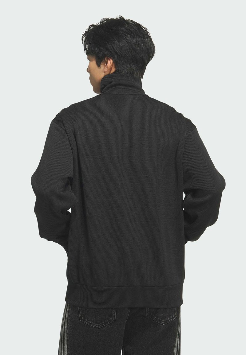 Man met kort donker haar die een zwarte coltrui en donkere spijkerbroek draagt, gezien van achter tegen een effen achtergrond.