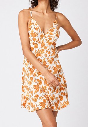 Mouwloze witte jurk met oranje bloemmotief, V-hals en dunne bandjes, gedragen door een persoon met één hand op de heup en de andere hand die de zoom van de jurk vasthoudt.