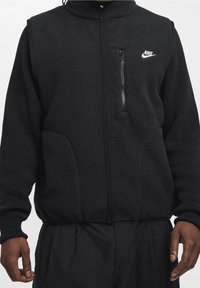 Svart Nike-väst med en texturerad fleeceframsida, släta stickade sidor, full dragkedja, hög krage och framficka med dragkedja.