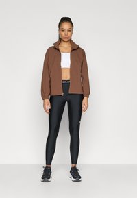 Καφέ ζακέτα με φερμουάρ πάνω από λευκό cropped τοπ, μαύροι κολάν με λογότυπο και μαύρα αθλητικά παπούτσια. Λείο ύφασμα, εφαρμοστή σχεδίαση και μακριά μανίκια.