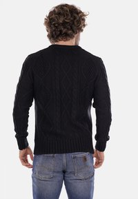 Maglione nero a maglia con un motivo testurizzato che presenta trecce e diamanti; polsini e orlo a costine. Indossato con jeans blu.