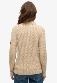 Beige kabelgebreide trui met een geribbelde kraag en manchetten. Opvallende patronen omvatten verhoogde gedraaide kabels. Draagt bij een spijkerbroek.