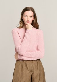 Hellrosa gestrickter Pullover mit weicher Textur, langen Ärmeln und gerippten Bündchen, kombiniert mit braunen, strukturierten, weit geschnittenen Hosen.