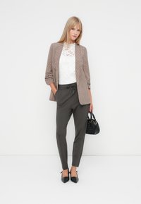Blazer à carreaux marron sur un top en dentelle blanche, associé à un pantalon vert foncé et des escarpins noirs à bouts pointus. Sac à main noir avec un détail texturé.