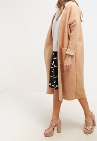 Cappotto lungo e aperto davanti in morbido tessuto beige con grandi tasche e maniche arrotolate; abbinato a una gonna a righe bianche e nere e sandali con piattaforma rosa.