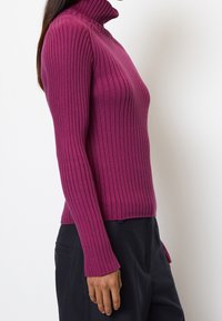 Fitted turtleneck sweater in deep magenta with ribbed texture, long sleeves, and a slightly cropped hem, paired with black pants.