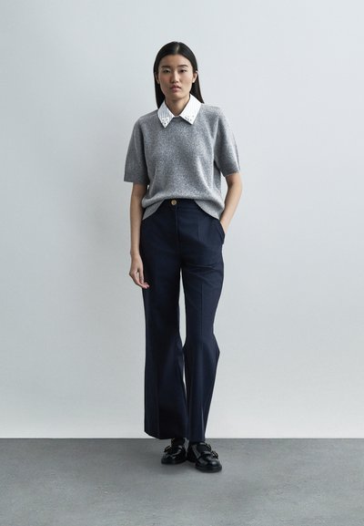 Jeune femme debout contre un mur uni, portant un pull gris à manches courtes sur une chemise blanche à col, un pantalon large bleu marine et des mocassins noirs.