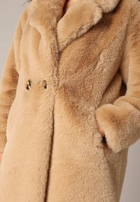 Abrigo de piel sintética beige con cuello de chal, diseño de doble botonadura con botones en tono dorado y textura suave.