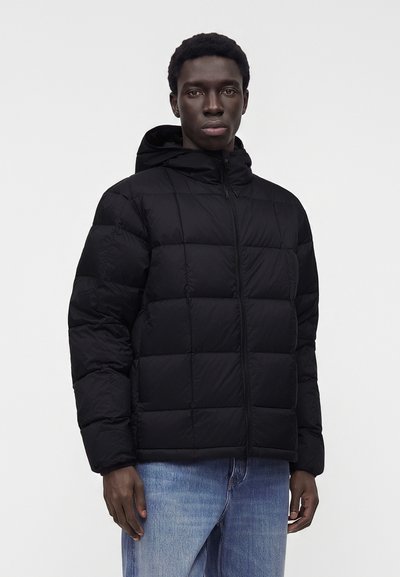 Homme portant une veste matelassée noire à capuche et un jean bleu, debout devant un fond clair uni.