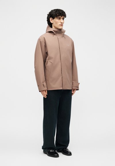 Giovane uomo con capelli ricci indossa una giacca con cappuccio marrone chiaro, pantaloni neri a gamba larga e scarpe nere su uno sfondo semplice.