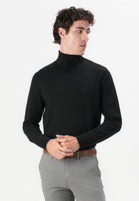 Crna turtleneck džemper u mekoj pletenini, s uskim krojem i dugim rukavima, s rebrastim manžetama, uparen s sivim hlačama.