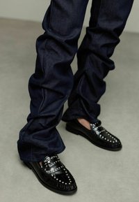 Zwarte loafers met zilveren studs, bovenwerk van textuurleer, gecombineerd met donkerdenim broek met een geplooid ontwerp en een gladde stofafwerking.