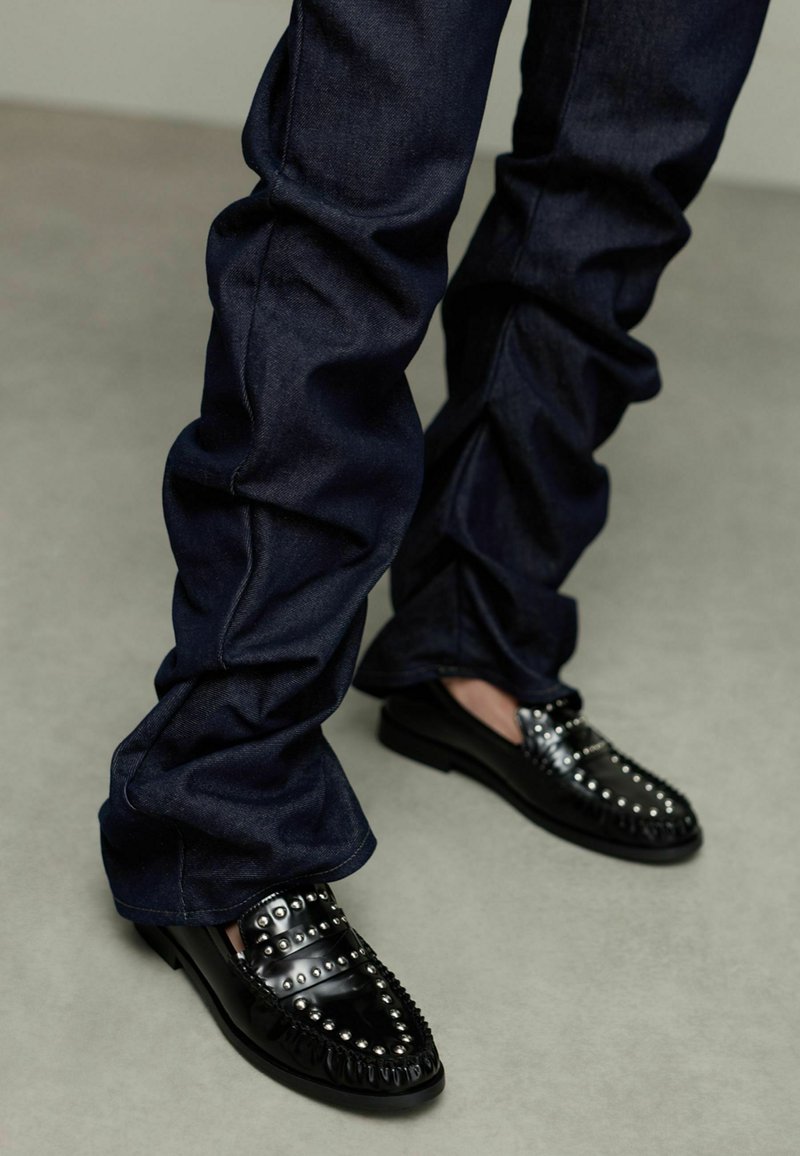 Zwarte loafers met zilveren studs, bovenwerk van textuurleer, gecombineerd met donkerdenim broek met een geplooid ontwerp en een gladde stofafwerking.