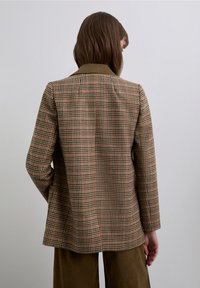 Blazer de cuadros marrón con un corte entallado, que presenta un cuello con muesca y una textura suave. La tela incluye sutiles acentos rojos y verdes.