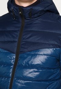 Marineblauwe pufferjas met een ritssluiting aan de voorkant, capuchon en een mix van matte en glanzende texturen met een chevronpatroon op de borst.