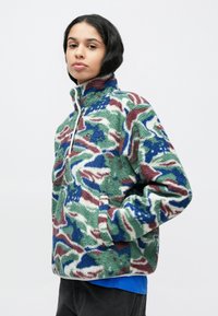 Φλις πουλόβερ με καμουφλάζ σε πράσινο, μπλε και μπορντούρα, με ψηλό γιακά και μπροστινό φερμουάρ μέχρι τη μέση. Μαλακή υφή με πλαϊνές τσέπες.