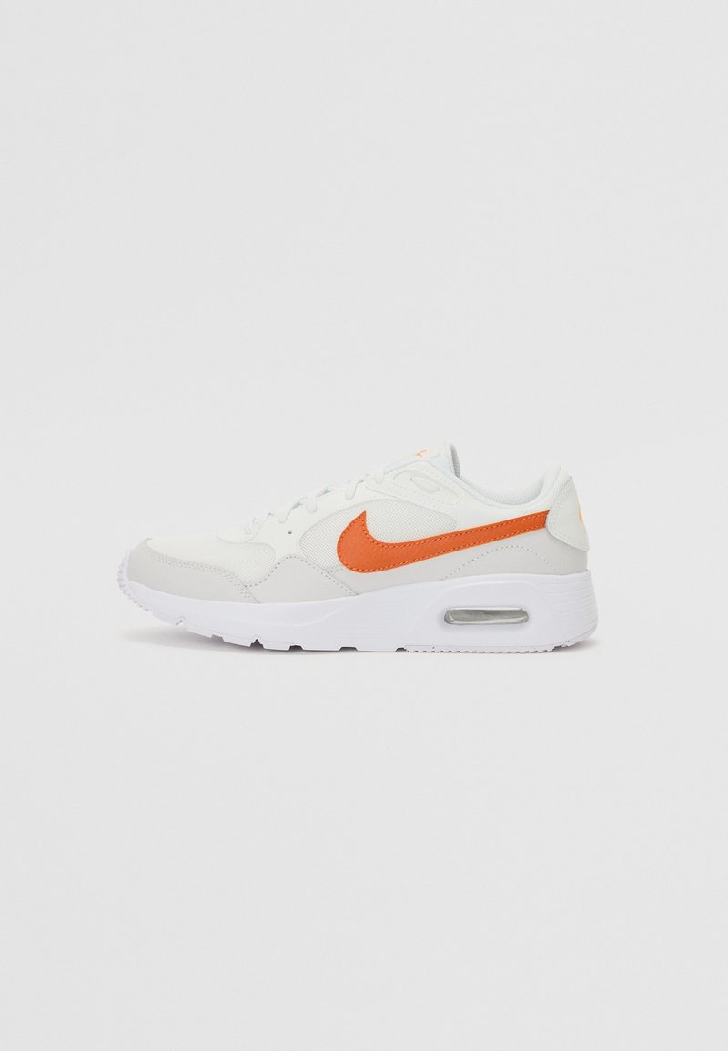 Zapatilla blanca con el logo de Nike en naranja, paneles lisos y texturizados, punta redonda y amortiguación visible en la suela.