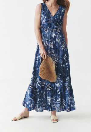 Femme portant une robe longue bleue sans manches avec des motifs de feuilles blanches et foncées, tenant un chapeau de soleil tressé et portant des sandales beige à enfiler.