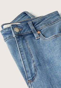 Jean en denim bleu plié montrant une poche avant, une boucle de ceinture, un bouton et un détail de rivet métallique sur fond clair.
