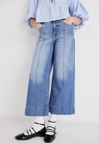 Jeans a vita alta e gamba larga di colore blu chiaro con finitura sbiadita, dotati di tasche laterali e dettagli cuciti, abbinati a ballerine nere e calzini bianchi.