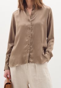 Brown silk blouse with a buttoned front, mandarin collar, and long sleeves. Soft texture and a relaxed fit, paired with light linen trousers.