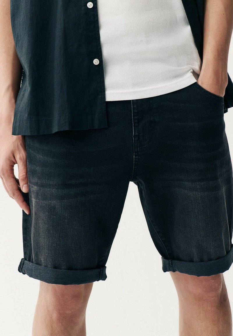 Temni jeans kratke hlače s zavihanimi robovi, z diskretno teksturo in prilegajočim krojem. V kombinaciji z belo rebrasto majico brez rokavov in odprto srajco z gumbki.