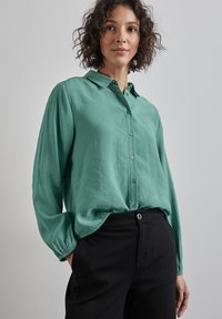 Chemise à manches longues, col en tissu vert doux, avec des boutons sur le devant, coupe décontractée, associée à un pantalon noir taille haute. Texture subtile.