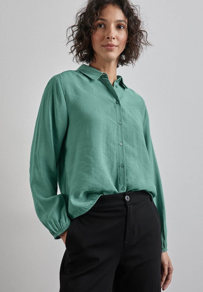 Chemise à manches longues, col en tissu vert doux, avec des boutons sur le devant, coupe décontractée, associée à un pantalon noir taille haute. Texture subtile.