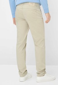 Pantaloni beige chiaro in cotone con vestibilità slim, dotati di tasche posteriori e logo ricamato sulla vita. Indossati con scarpe da ginnastica bianche.