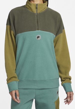 Femme portant un pull polaire color-block avec demi-fermeture éclair en bleu sarcelle, vert olive et jaune moutarde, associé à un pantalon bleu sarcelle.