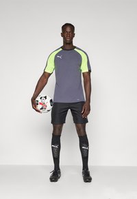 Grå kortermet sportskjorte med neon grønne detaljer, svarte shorts, svarte sokker med logo, og svarte fotballsko som holder en mønstret fotball.