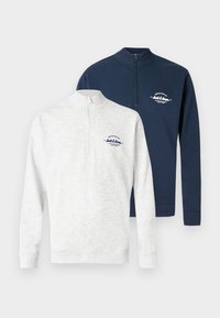 Δύο πουλόβερ με φερμουάρ μέχρι τη μέση: το ένα ανοιχτό γκρι με υφή και ναυτικές λεπτομέρειες· το άλλο ολόκληρο ναυτικό μπλε με ομαλό ύφασμα, και τα δύο με μικρά λογότυπα.