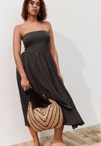 Vestito nero senza spalline con gonna fluida e arricciata in tessuto leggero e testurizzato. Modella che tiene una borsa intrecciata di paglia a forma di cuore.