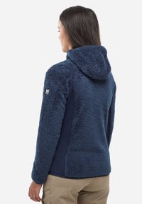 Kobieta stojąca tyłem, ubrana w niebieską polarową kurtkę z kapturem i beżowe spodnie na białym tle.