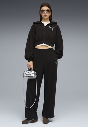 Vrouw draagt een zwarte Puma-trainingspak met een korte rits-hoodie, wijde broek, bruine schoenen, getinte zonnebril en houdt een kleine zilveren Puma-handtas vast.