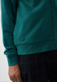 Sudadera azul verdosa con cremallera, que presenta un dobladillo acanalado, un tejido texturizado y un pequeño logotipo en relieve en el lado izquierdo cerca del dobladillo. Combinada con pantalones de cuadros.
