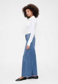 Bel dolg rokav bele bluze, kombinirane z visokim pasom in širokimi modrimi kavbojkami. Model nosi črne balerinke. Enostavna zasnova, brez vidnih vzorcev ali okraskov.