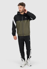Eingehüllte Jacke in Schwarz und Olivgrün mit weißen Akzenten. Kombiniert mit schwarzen Hosen mit seitlichen Streifen und Logo. Weicher, glatter Stoff.
