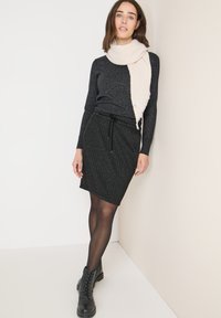 Zwarte gebreide trui, grijze gestructureerde kokerrok met trekkoord, doorschijnende zwarte panty's en zwarte veterschoenen. Crème sjaal over de trui gedrapeerd.