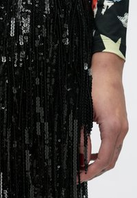 Tela negra con lentejuelas y detalles de flecos, que muestra una textura brillante y un diseño en capas. Una mano con esmalte de uñas oscuro sostiene el material.