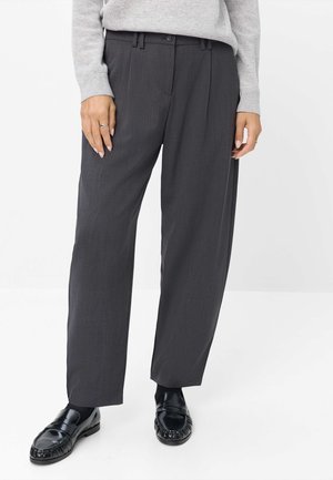Personne portant un pantalon large gris foncé, un pull gris clair, des chaussettes noires et des mocassins en cuir noirs debout sur un fond blanc.