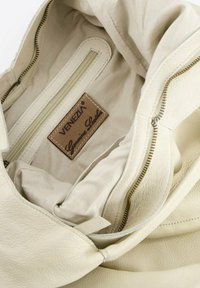 Kremowa skórzana torba wewnątrz z beżowym wykończeniem. Zawiera kieszonkę na zamek oraz brązową metkę z napisem "VENEZIA Genuine Leather."