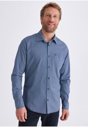 Mann mit hellbraunem Haar und Bart, der ein blau gemustertes Langarmhemd mit Knöpfen und dunkle Hose trägt, vor weißem Hintergrund steht.