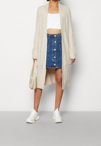 Hellbeige gestrickte Strickjacke, lange Ärmel, offene Vorderseite. Kombiniert mit einem weißen Crop-Top und einem Jeansrock mit Knopfdetails. Weiße Turnschuhe.