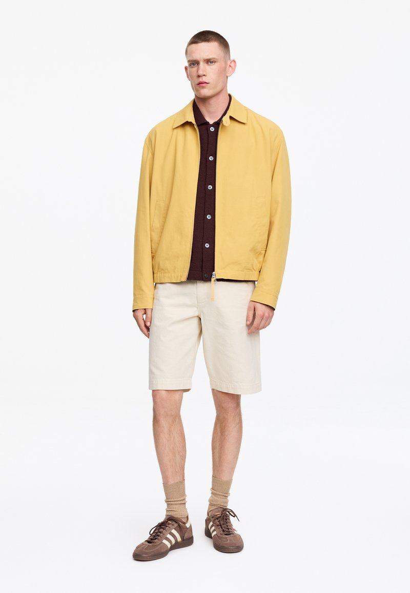 Jeune homme debout, portant une veste jaune zippée, une chemise marron foncé boutonnée, un short beige, des chaussettes beige et des baskets marron avec des rayures blanches.