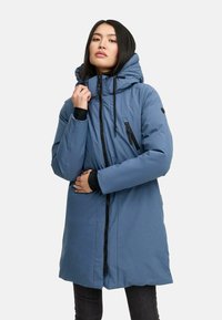 Donna che indossa un cappotto invernale blu imbottito con cappuccio, in piedi e tiene la zip vicino al collo contro uno sfondo chiaro.