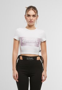 Valkoinen lyhyt crop-top, jossa on keskellä vaaleanpunainen paljettilogot. Lyhyet hihat, pyöreä kaula-aukko, yhdistetty mustiin korkeavyötäröisiin housuihin, joissa on logovyötärö.