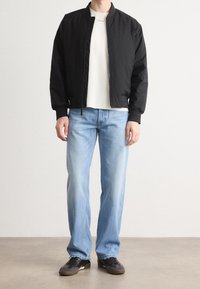 Chaqueta bomber negra con cremallera en el frente, llevada sobre una camiseta blanca, combinada con jeans de corte relajado azul claro y zapatillas negras.